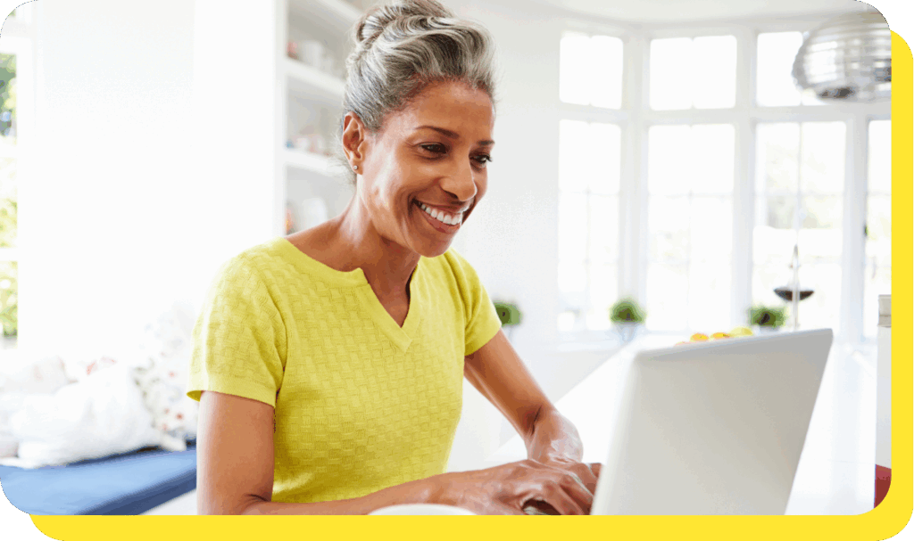 A woman smiling warmly while seated at a desk, using her laptop to explore a credit union loan calculator.