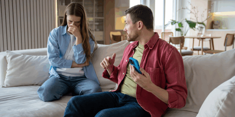 Man sat on sofa hiding phone from partner, who is visibly concerned.