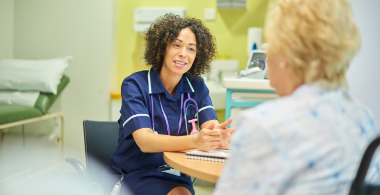 An NHS professional in a blue tunic sitting at a table and speaking with a patient in a bright clinic room.